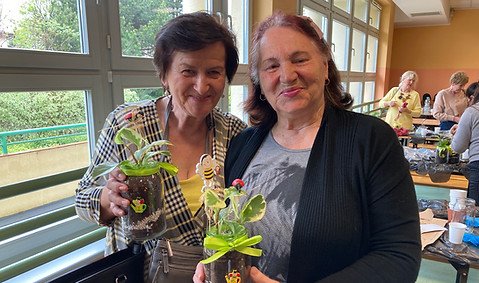 Zdjęcie z zajęć praktycznych z Gdańskiej Akademii Seniora - filia Osowa. Zdjęcie przedstawia dwie starsze, uśmiechnięte kobiety, które trzymają doniczki z własnymi kwiatami.