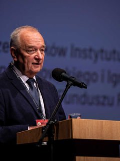 Facet w starszym wieku w garniturze, stoi przy mównicy zwrócony do mikrofonu. 