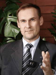 Uśmiechnięty pan w średnim wieku. Siedzi w fotelu,jego lewa ręka gestykuluje. Ma na sobie czarny garnitur z krawatem w psaki i białą koszulą. 