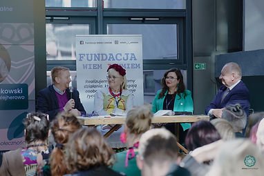 Zdjęcie z panelu dyskusyjnego z konferencji "Etnopomorze2022 ". Przedstawia cztery osoby. Dwie kobiety siedzące w środku i dwóch mężczyzn na zewnątrz. Mężczyzna o lewej trzyma mikrofon, a reszta się uśmiecha w jego kierunku. 