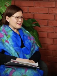 Siedząca pani w kasztanowych krótkich do ramion włosach, owinięta w kolorowy szal z dominującym niebieskim kolorem, nosi okulary korekcyjne z czarną cienką oprawką, a na kolanach ma położoną teczkę.