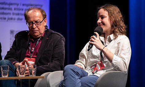 Zdjęcie z konfencji "Talnet dla wspólnoty" z panelu dyskusyjnego. Po prawej stronie siedzi uśmiechnięta licealistka, trzymająca mikrofon, a po lewej starszy pan z założonymi rękami, który w skupieniu słucha dziewczyny.