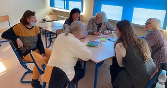 Zdjęcie przedstawia 5 kobiet w starszym wieku i 1 z młodym. Kobiety siedza w kole przy dwóch złączonych ławkach szkolnych i rozmawiają.