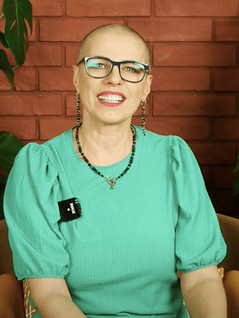 Łysa kobieta w średnim wieku w zielonej bluzce, z koralikami na szyi, z okularami korekcyjnymi na nosie. 