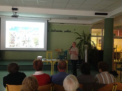 Zdjęcie z wykładu. Zdjęcie przedstawia starszą kobietę trzymającą mikrofon, która stoi zwrócona przedem do uczestników, a tyłem do projektora.