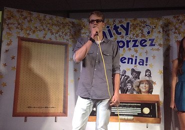 Zdjęcie chłopaka który śpiewa do mikrofonu a za nim jest plakat okładki płyty "Hity przez pokolenia"